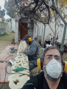 שני אומנים במסכות אבק ואוזניות מגן מלטשים פסי גילוף מעץ בחצר הסדנה