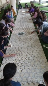 בנות יושבות לאורך יריעת סבכה לבנה על דשא סינתטי ומרכיבות את הדוגמה