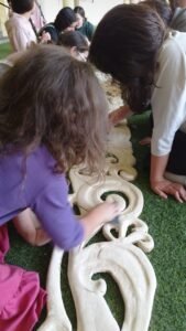 בנות מלטשות אלמנט עץ מגולף בדוגמת עלים מסולסלים על דשא סינתטי