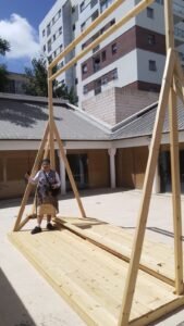 מסגרת נול עץ ענקית לאריגת הפרוכת מוקמת בחצר, ואישה עומדת לצידה