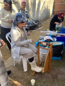 נשים בחצר סביב גלגל טווייה ומכלים של צמר צבוע