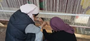 שתי אורגות קושרות חוטי שתי על הנול, מאחוריהן דגם הפרוכת הצבעוני