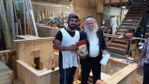 בעל המלאכה לצד רב מזוקן האוחז ספר, בנגרייה בין חלקי דגם בית המקדש השלישי