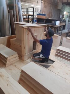 האמן כורע בנגרייה ומרכיב חלק עץ אורן גדול מדגם בית המקדש השלישי בשלבי בנייה