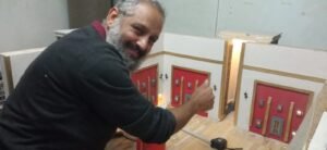 האמן מחייך עם סרט מדידה ליד אגף צבוע בדגם — קירות לבנים ודלתות אדומות עם משקופים מוזהבים